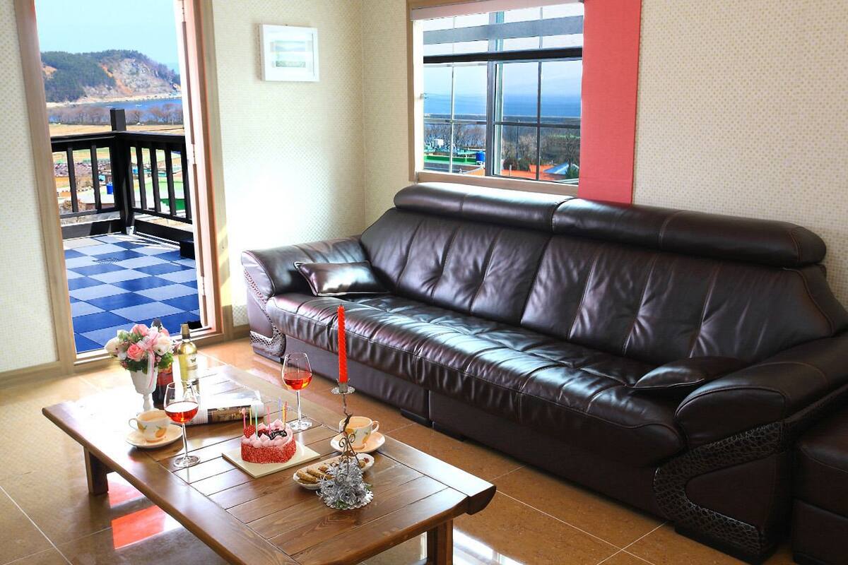 Un salon avec un canapé en cuir foncé, une table basse en bois sur laquelle sont posés un gâteau et des boissons, et une vue sur une colline depuis un balcon.