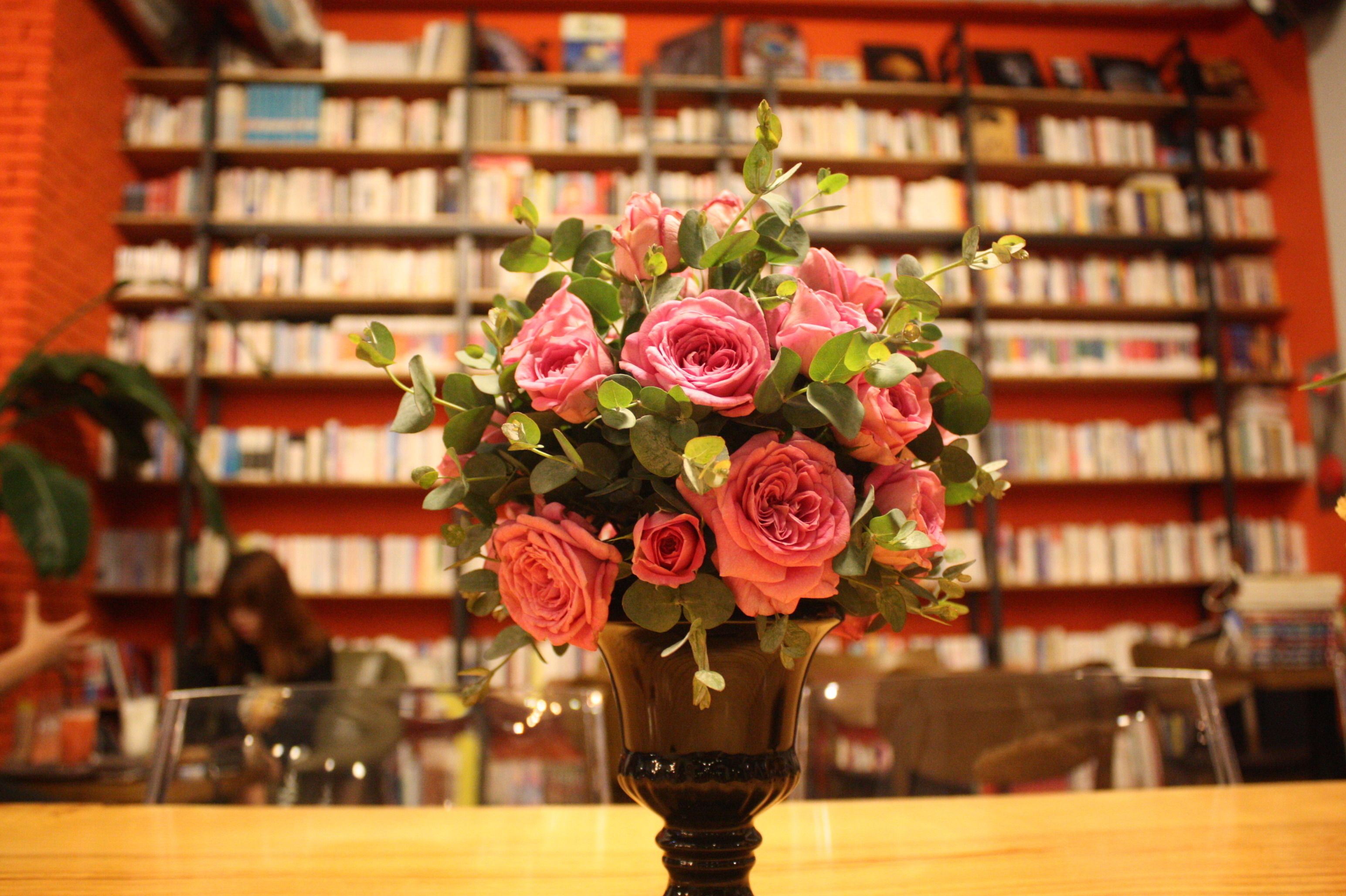 A bouquet of pink roses in a dark vase on a wooden table in a library setting with bookshelves in the background.