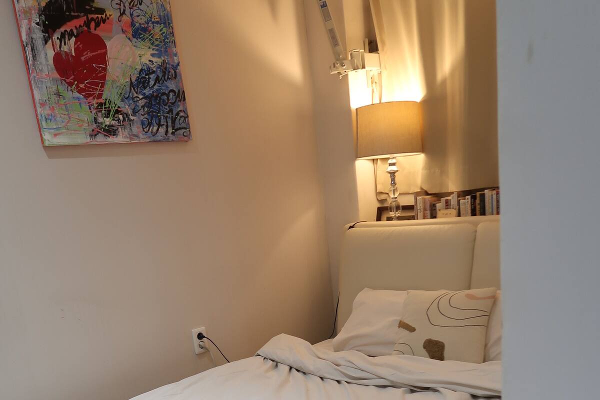 A small, neatly made bed with a white comforter and a note on it. There is a white bedside table with a lamp and a few books. A colorful abstract painting hangs on the wall above the bed.