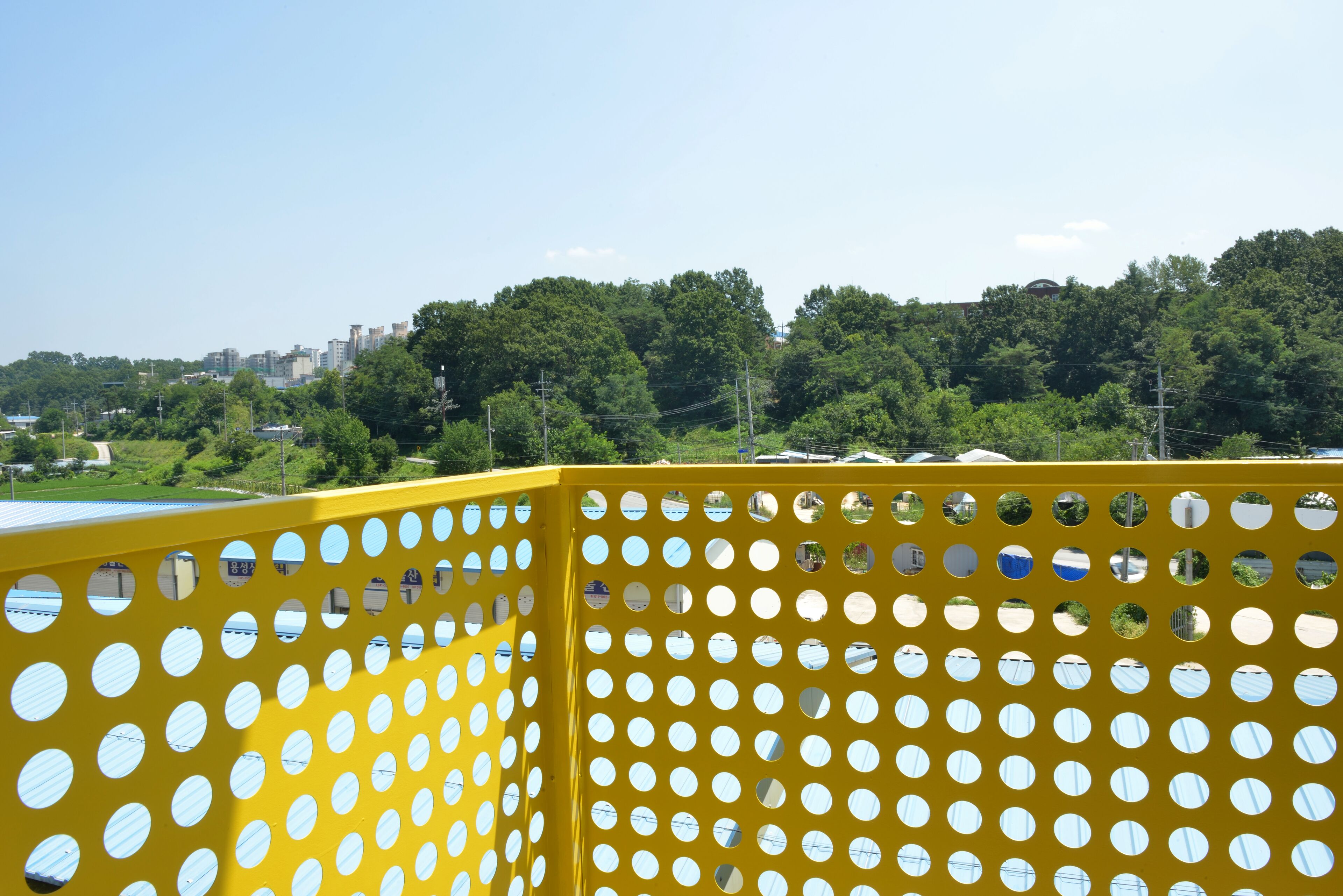 Una valla de metal amarillo con recortes circulares, un paisaje verde con árboles y edificios al fondo.