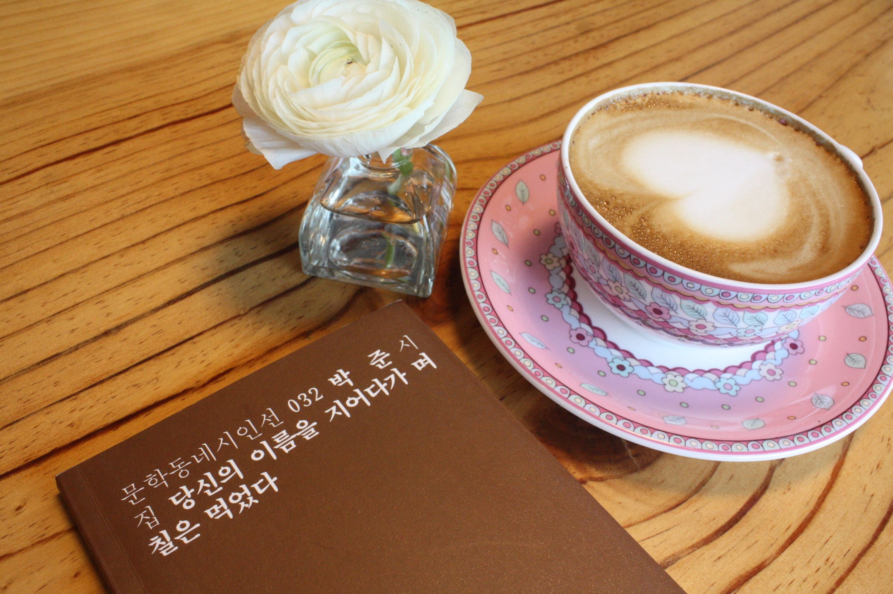 A cup of coffee with latte art on a pink saucer, a small glass vase with a white flower, and a brown booklet with Korean text.