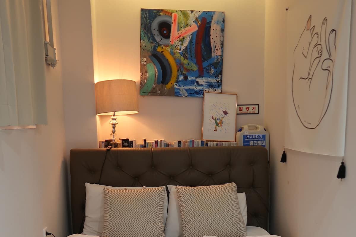 A neatly made bed with a headboard, a bedside lamp, and a colorful abstract painting above it.