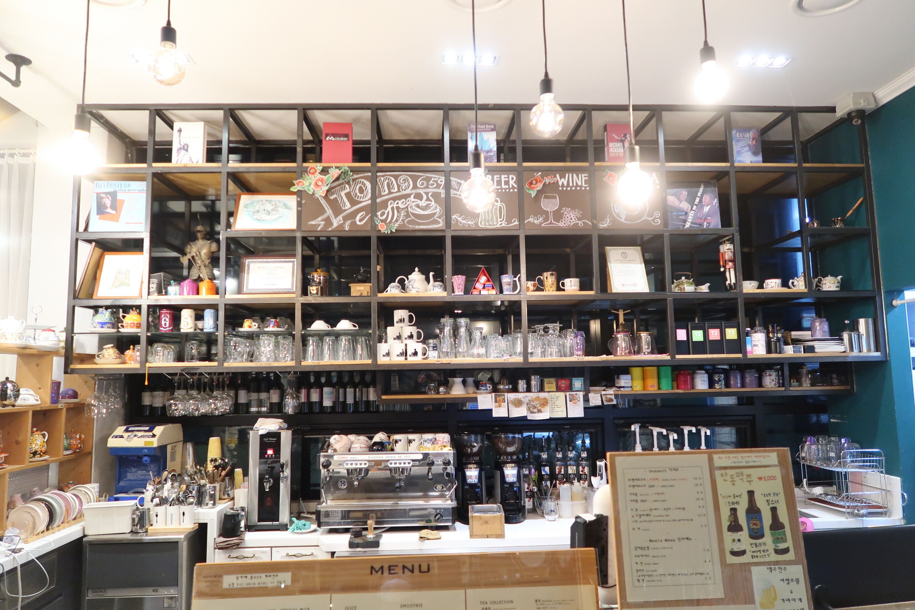 Un café con estantes llenos de tazas, vasos y vajillas de té. Se observa una máquina de café y un panel con la carta.