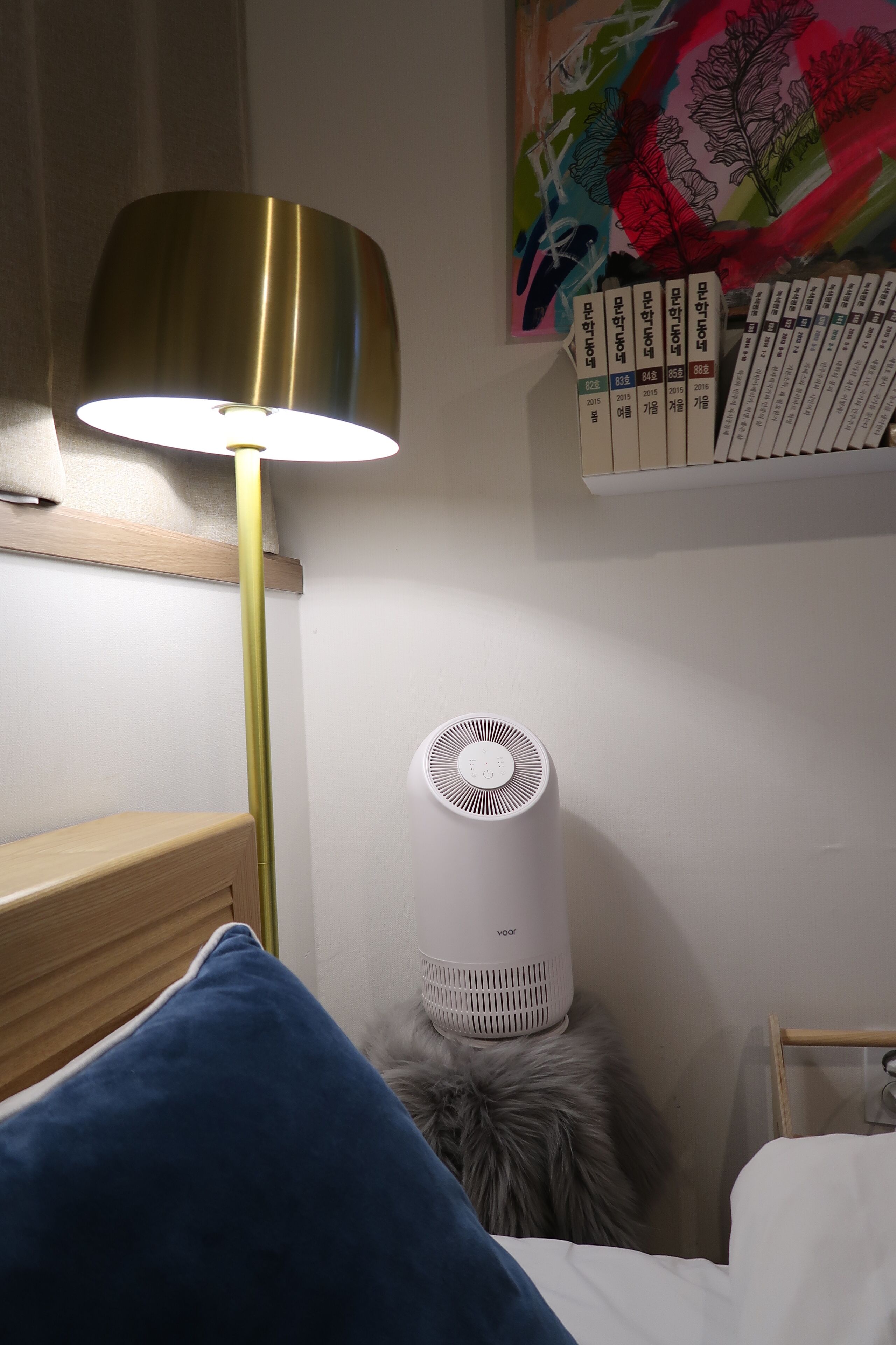 Een slaapkamer met een bed, een blauw kussen, een lamp en een witte luchtreiniger.