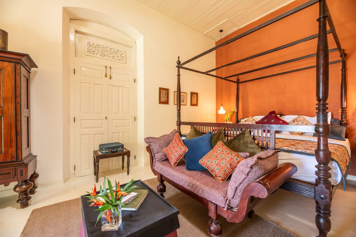 A four-poster bed with a wooden frame, a sofa with multiple cushions, a small table with a vase of flowers, and a wooden cabinet.
