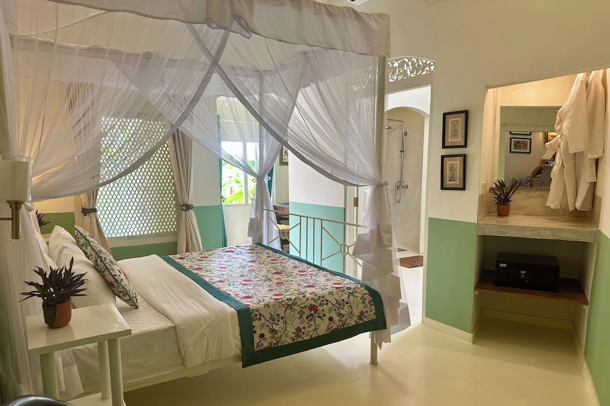 A bedroom with a canopy bed, a small table with a plant, a shower area, and a mirror with a towel rack.