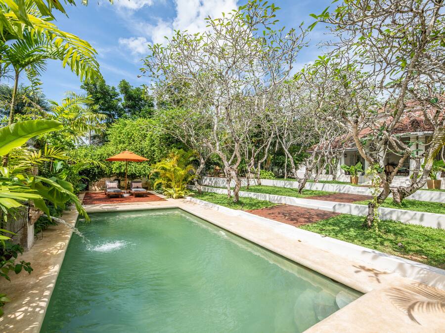 Una piscina rectangular con un cielo azul despejado, rodeada de abundante vegetación y una cerca blanca.