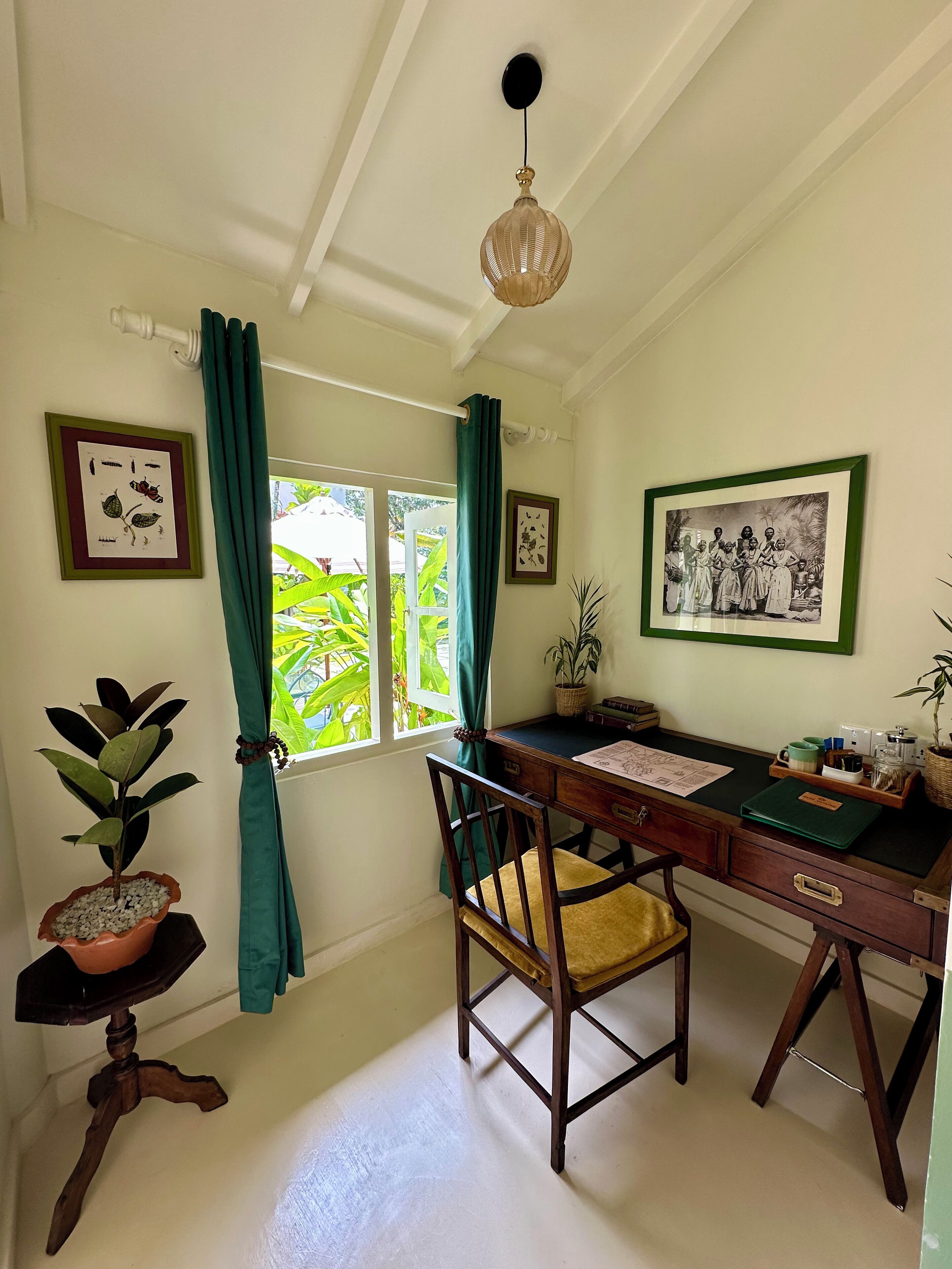 Quarto com escrivaninha de madeira, uma cadeira, planta em vaso, impressão botânica emoldurada, luminária pendente e janela com cortinas.