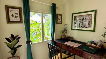 Quarto com escrivaninha de madeira, uma cadeira, planta em vaso, impressão botânica emoldurada, luminária pendente e janela com cortinas.