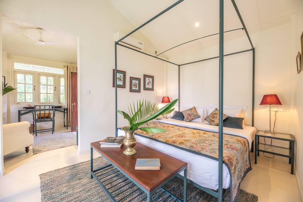 A bedroom with a four-poster bed, a bedside table with a lamp, and a view of the dining area.