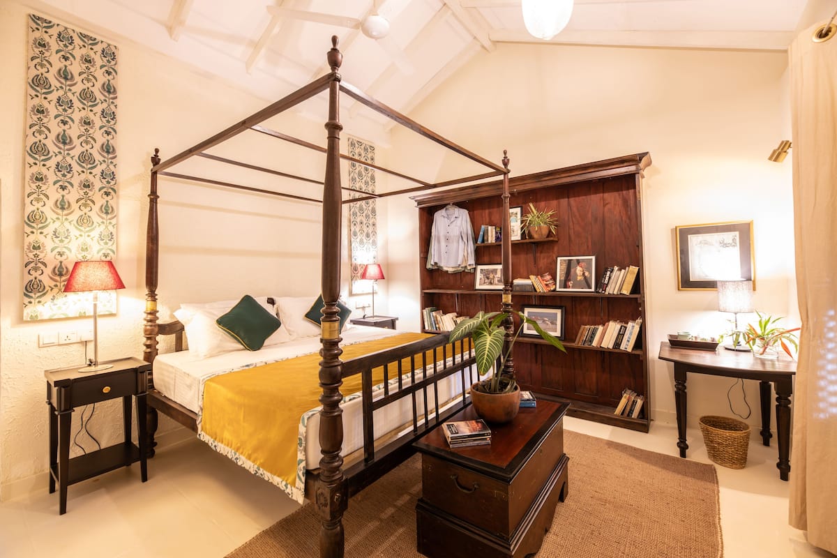 A four-poster bed with a wooden frame, a bedside table with a lamp, a bookshelf, and a small table with a plant.