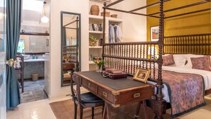 Quarto com cama de dossel, escrivaninha de madeira com gavetas, cadeira, estante e banheiro visível por uma porta aberta.