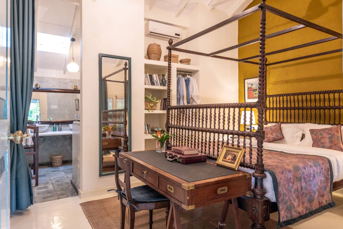 A bedroom with a four-poster bed, a wooden desk with drawers, a chair, a bookshelf, and a bathroom visible through an open door.