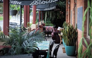 Un café al aire libre con mesas y sillas, plantas en macetas y un muro de ladrillo.