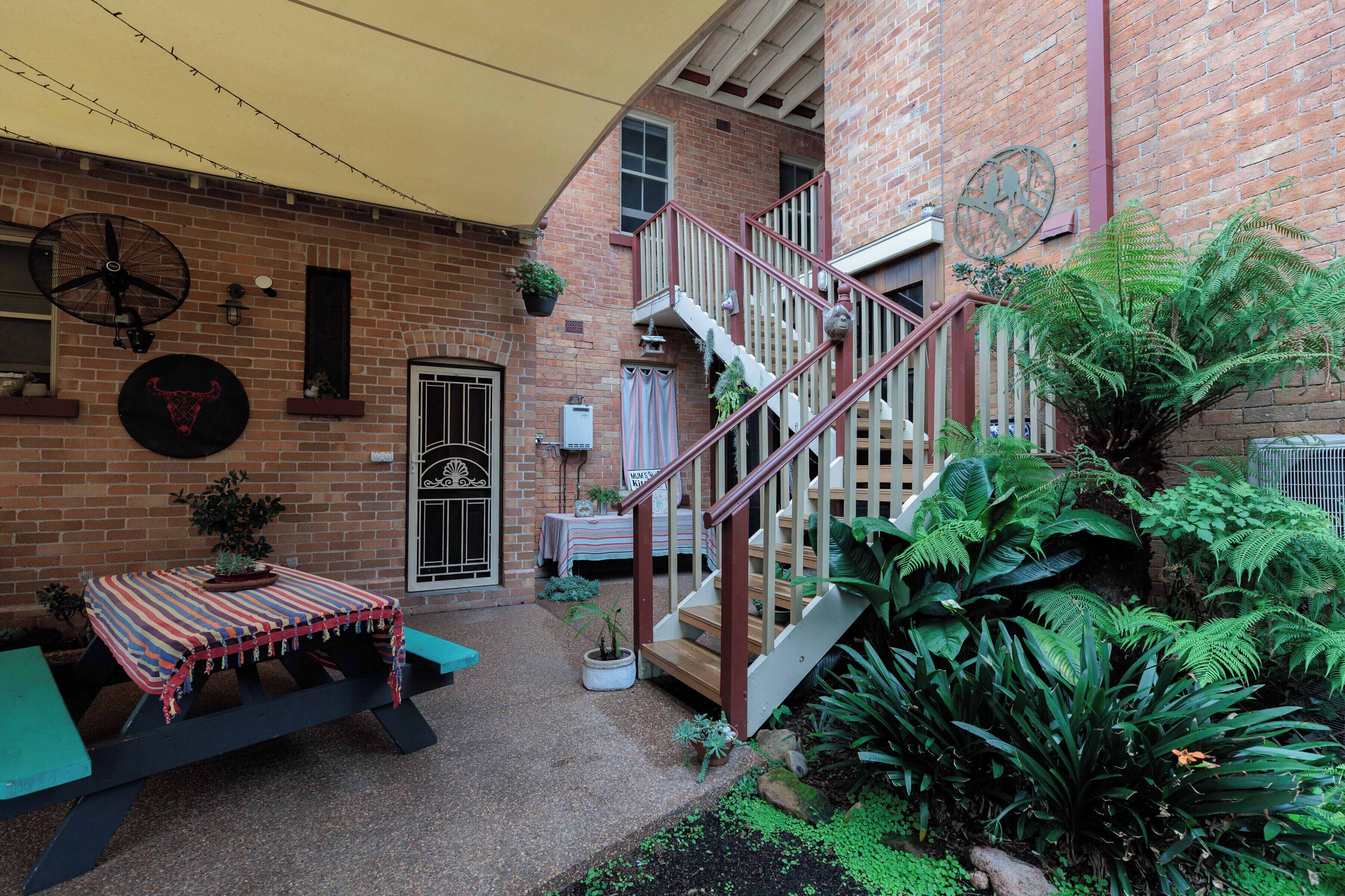 Un patio con un edificio de ladrillo, una escalera de madera, una mesa con mantel colorido y diversas plantas en macetas.