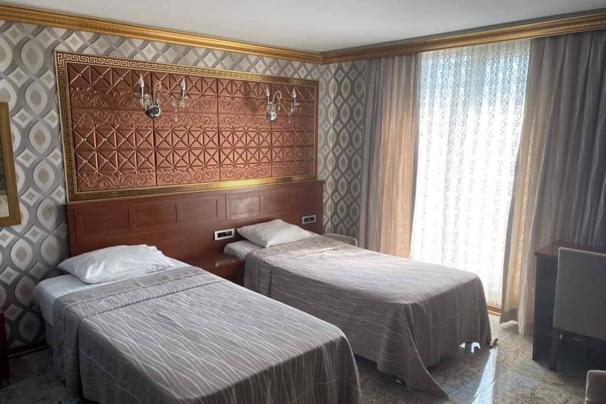 A hotel room with two single beds, a wooden headboard with intricate patterns, a small bedside table, and a large window with curtains.