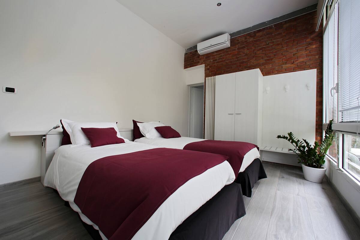 A modern bedroom with two beds, a potted plant, and a large window.