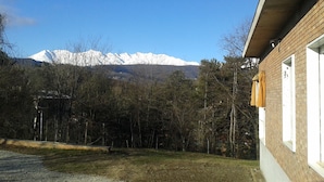 Un bâtiment à l’extérieur en briques, avec une fenêtre ornée d’un rideau. Au fond, on aperçoit des montagnes enneigées et une zone boisée.