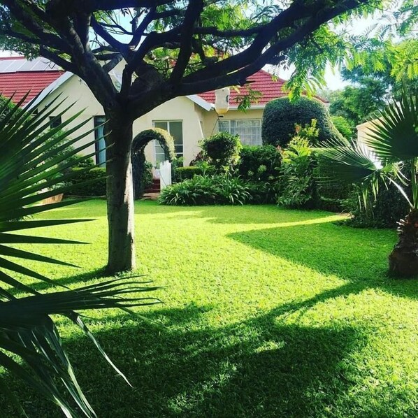 Un jardín cuidado con césped, un árbol y una casa con techo rojo al fondo.
