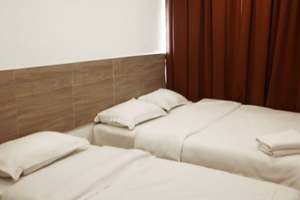 A hotel room with two beds, each with white bedding and pillows, a wooden headboard, and a window with brown curtains.