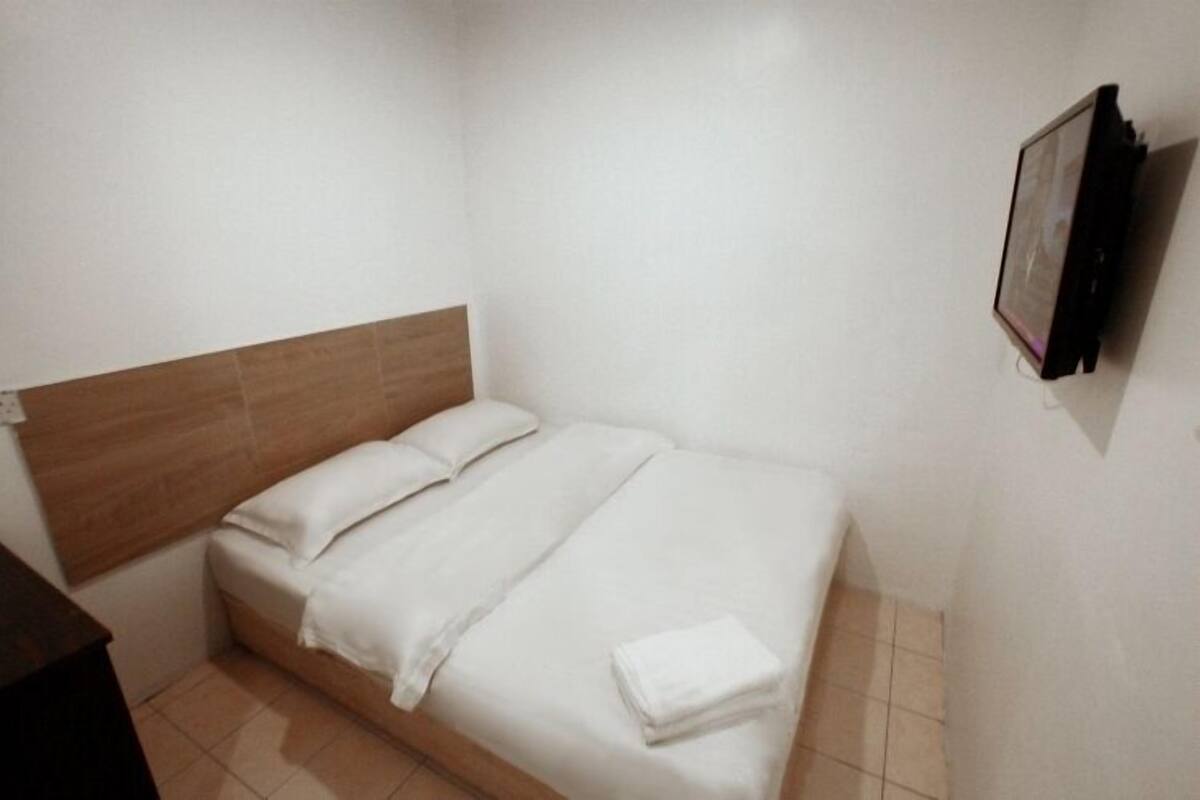 A small, neatly made bed with white linens in a compact room with a wooden headboard and a wall-mounted TV.