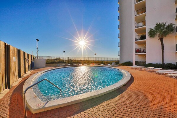 Una piscina con el sol reflejado en el agua, rodeada de sillones de descanso y un camino de ladrillo.