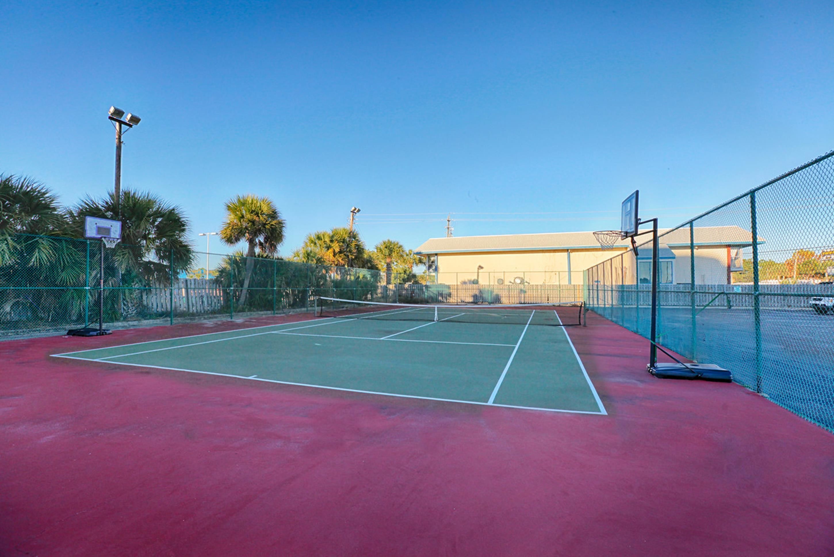 Un court de tennis en plein air, doté d’un filet et entouré d’une clôture grillagée, avec des palmiers en arrière-plan.