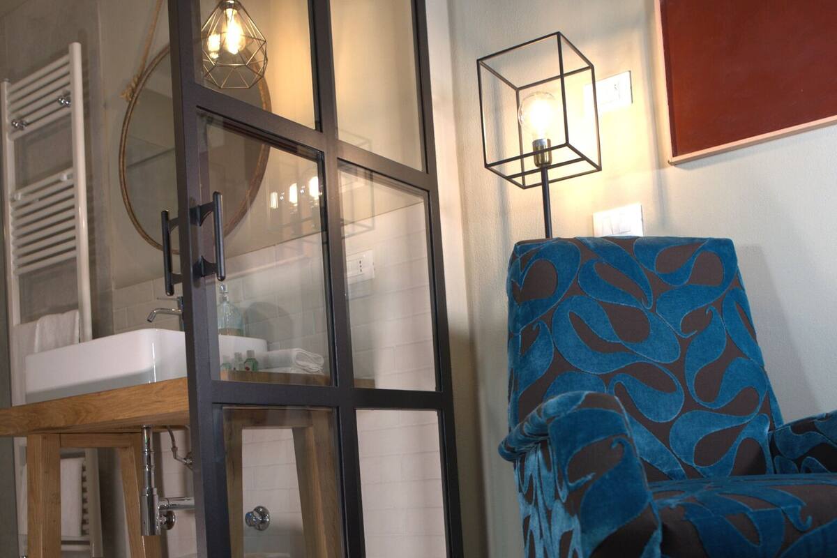 Une pièce avec une coiffeuse en bois, une chaise bleue à motifs, un tapis coloré et une porte en verre avec des panneaux dépolis.