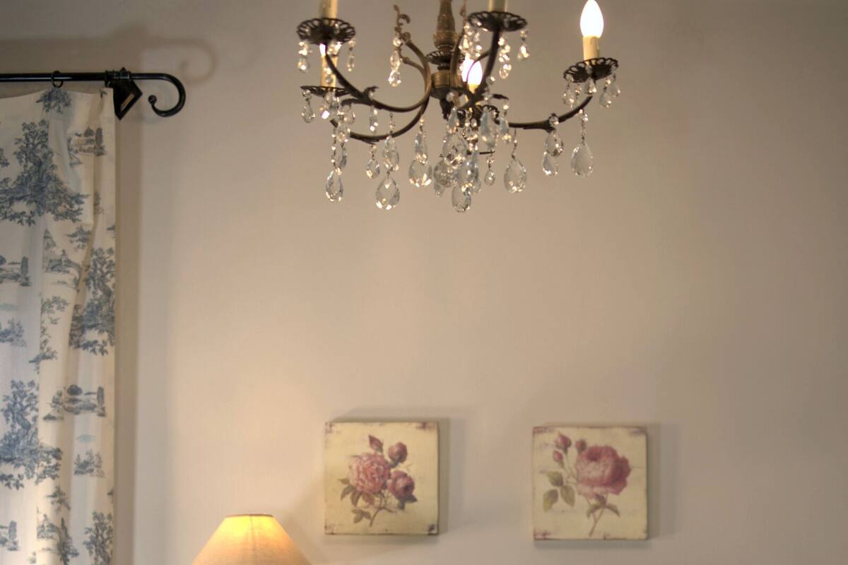Une chambre avec une commode bleue, un lit avec une literie blanche, un lustre et des œuvres d’art florales accrochées au mur.