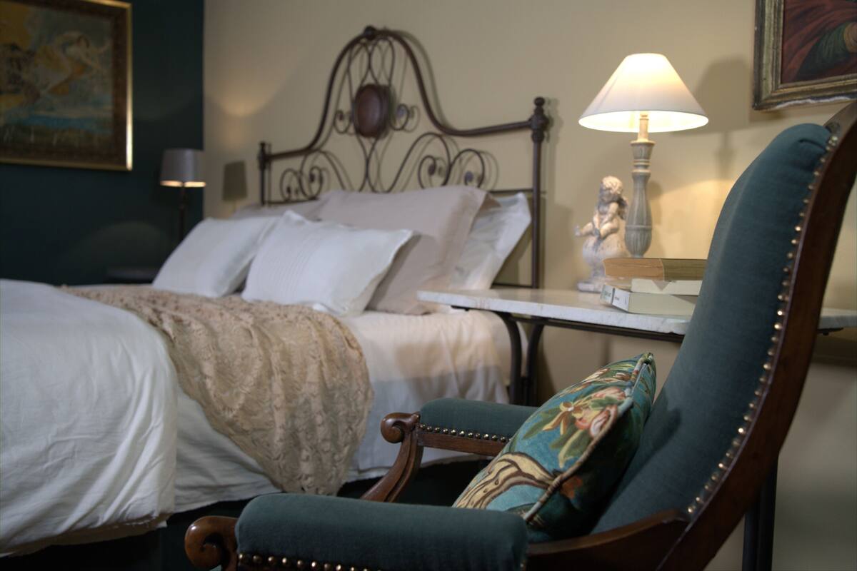 Un lit bien fait, avec du linge de lit blanc et une tête de lit décorative. Une chaise en bois, agrémentée d’un coussin à motifs floraux, est placée devant le lit.