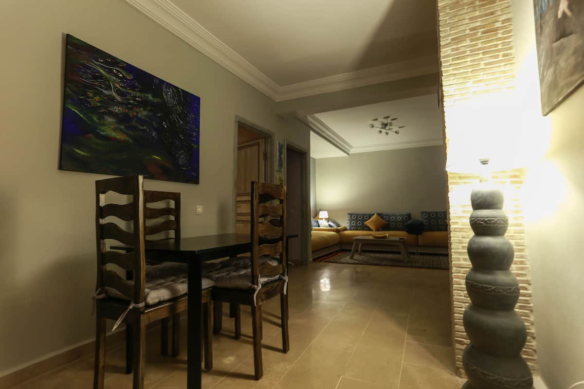 A dining area with a table and chairs, a painting on the wall, and a living room in the background with a sofa and lamp.
