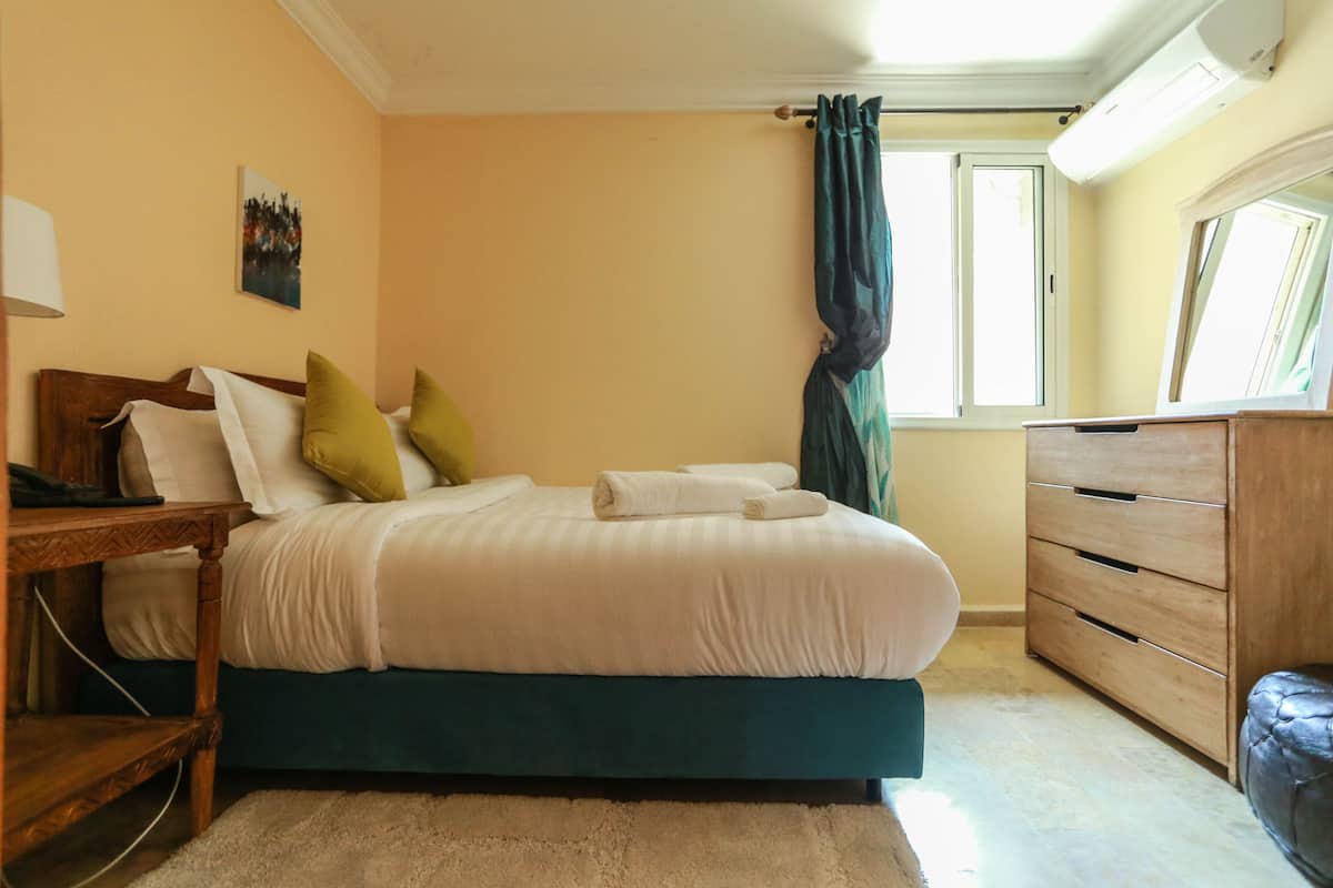 A bedroom with a bed, a dresser, a nightstand, a lamp, and a window with curtains.