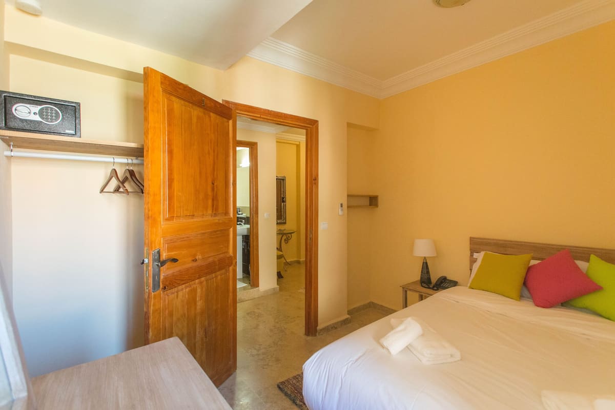 A hotel room with a bed, a wooden door, a nightstand, and a bathroom visible through an open door.