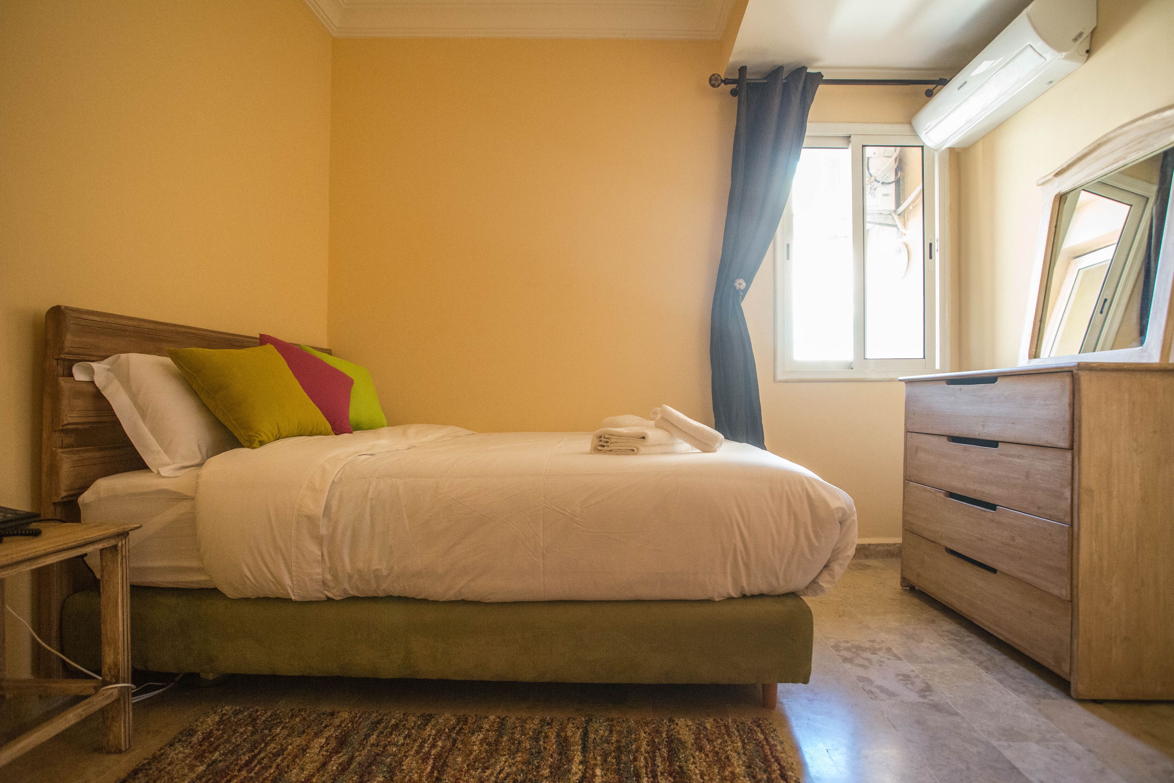 A neatly made bed with a green mattress, a wooden nightstand with a phone, a wooden dresser, and a window with a curtain.