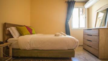 A neatly made bed with a green mattress, a wooden nightstand with a phone, a wooden dresser, and a window with a curtain.