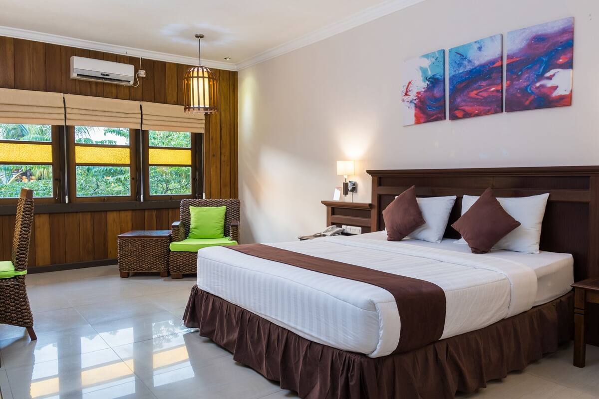 A hotel room with a large bed, a wooden headboard, a nightstand, a lamp, and a window with a view of greenery.