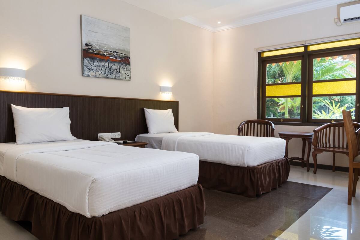 A hotel room with two beds, a desk, and a chair. There is a window with a view of greenery outside.