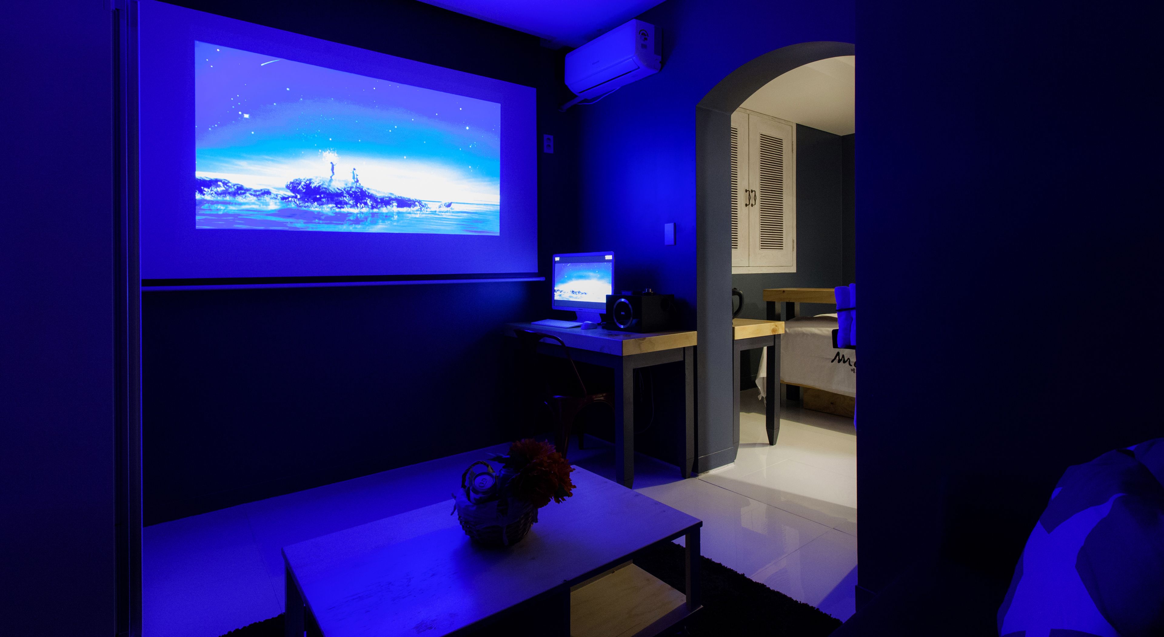 A room with a large screen displaying a scenic image, a desk with a computer, and a bed with a blue blanket.