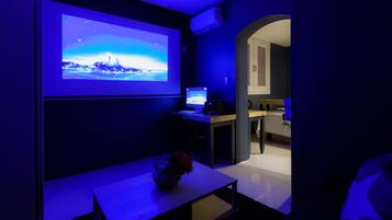 A room with a large screen displaying a scenic image, a desk with a computer, and a bed with a blue blanket.