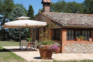 A rustic stone building with a tiled roof, a large potted plant with purple flowers, and a white umbrella.