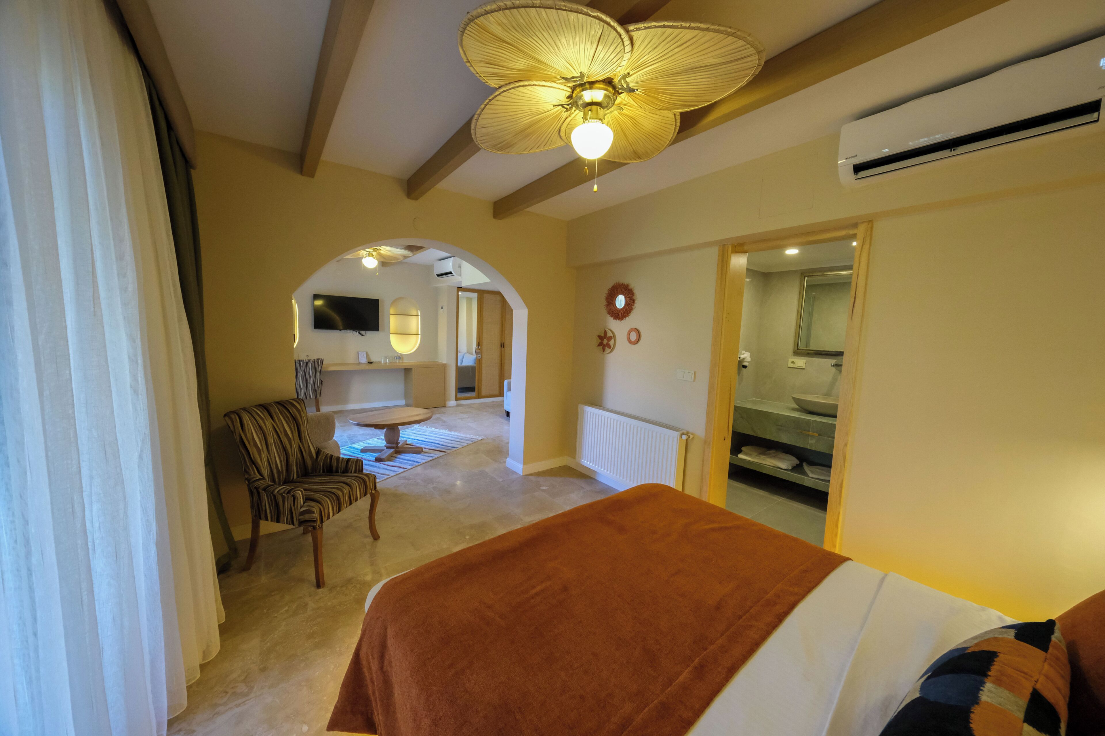 A hotel room with a bed, a chair, a table, a ceiling fan, and a view of the bathroom.