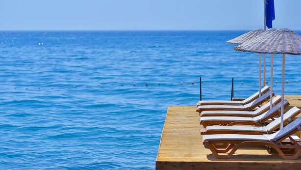Um deck de madeira com espreguiçadeiras e um guarda-sol azul, com vista para o mar calmo.