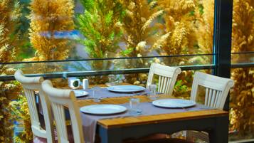 Mesa de jantar posta com pratos, copos e talheres, com vista para plantas altas e douradas através de uma janela.