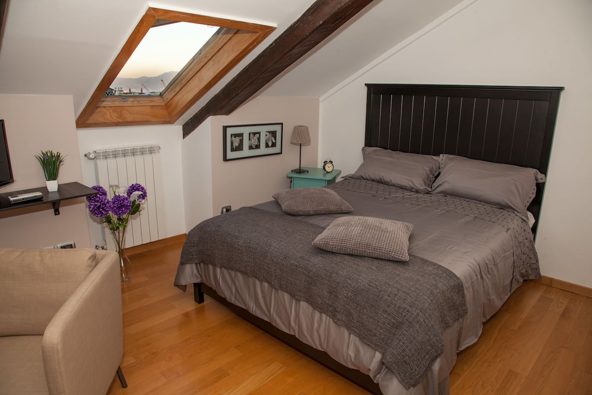 Quarto com teto inclinado, cama com cabeceira escura, mesa de cabeceira com abajur, quadro na parede e vista pelo claraboia.