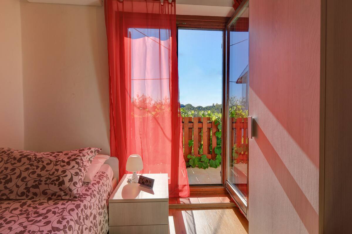 A room with a bed, a nightstand with a lamp, and a view of a wooden fence and greenery outside.