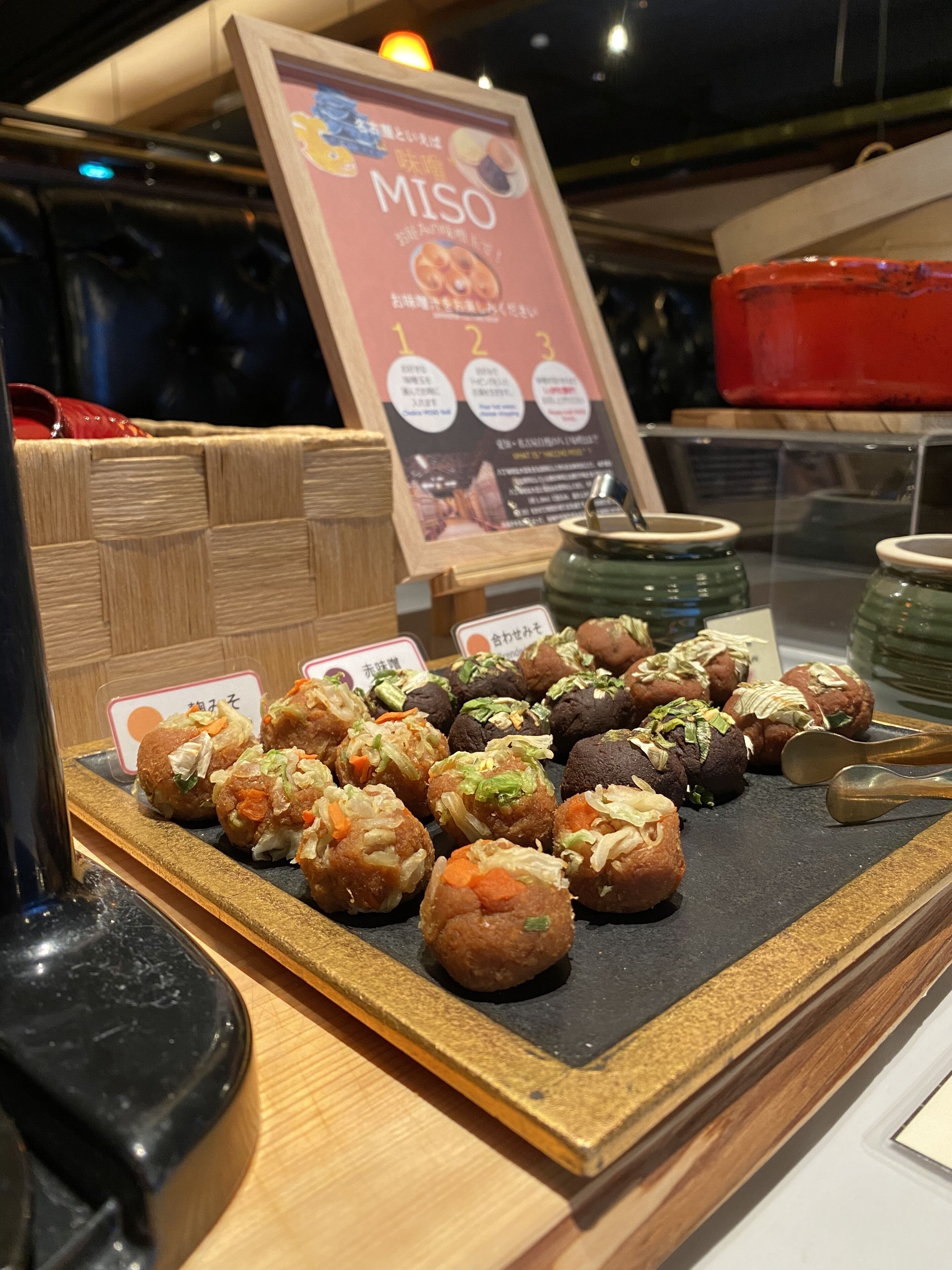 A display of various food items, including Miso and other snacks, arranged on a wooden counter with a sign above.