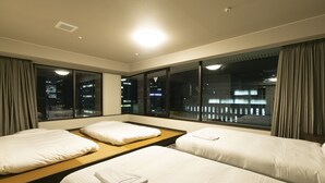 Habitación de hotel con tres camas, ventanas amplias y vista nocturna de la ciudad.