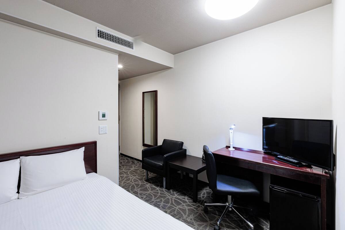 A hotel room with a bed, a desk with a TV, a chair, and a small table.
