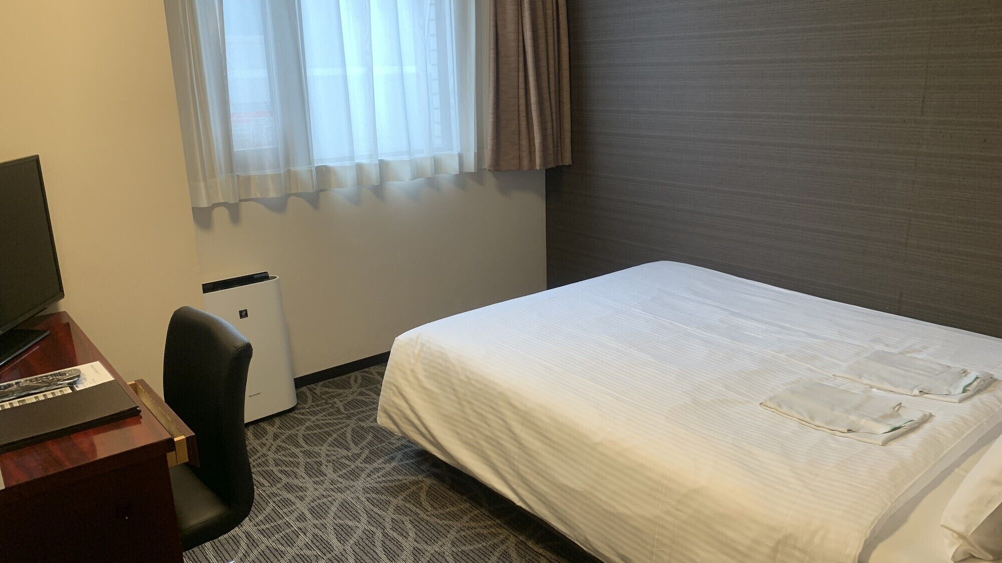 Een hotelkamer met een bed, een bureau met computer, een stoel en een raam met gordijnen.