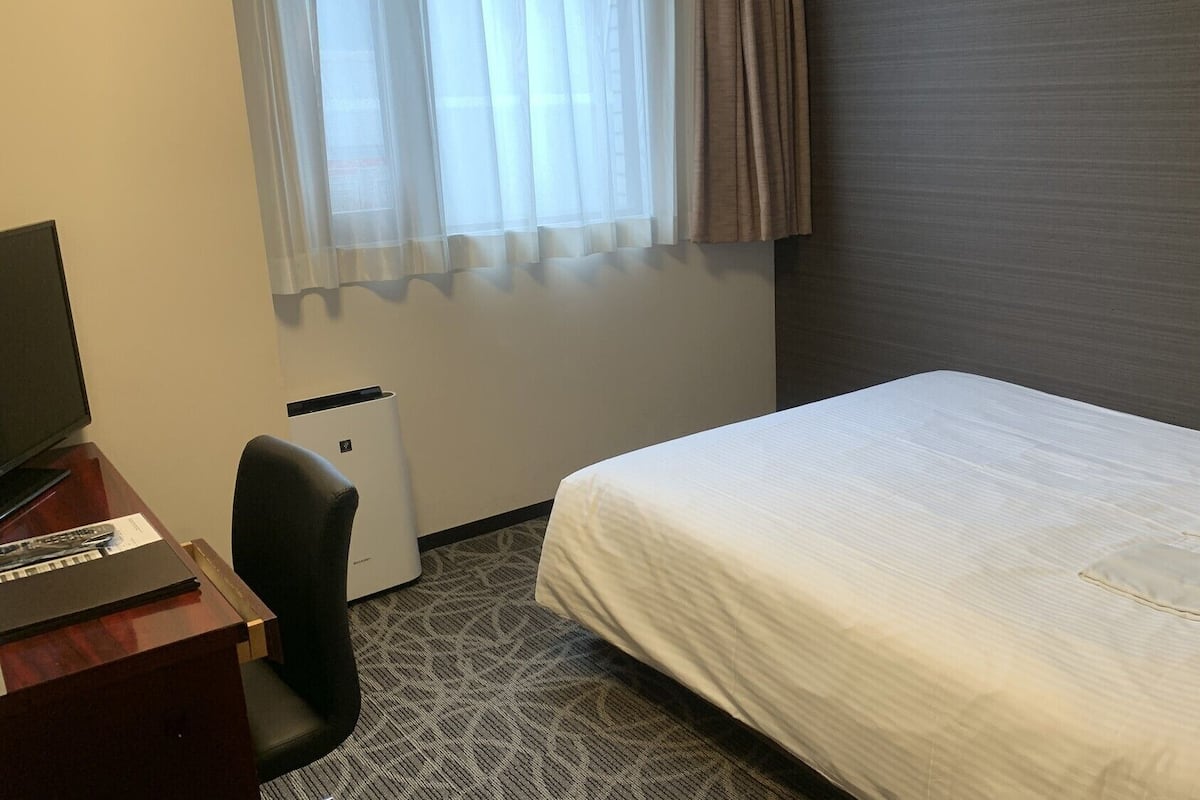 Une chambre d’hôtel avec un lit, un bureau avec un ordinateur, une chaise et une fenêtre avec des rideaux.